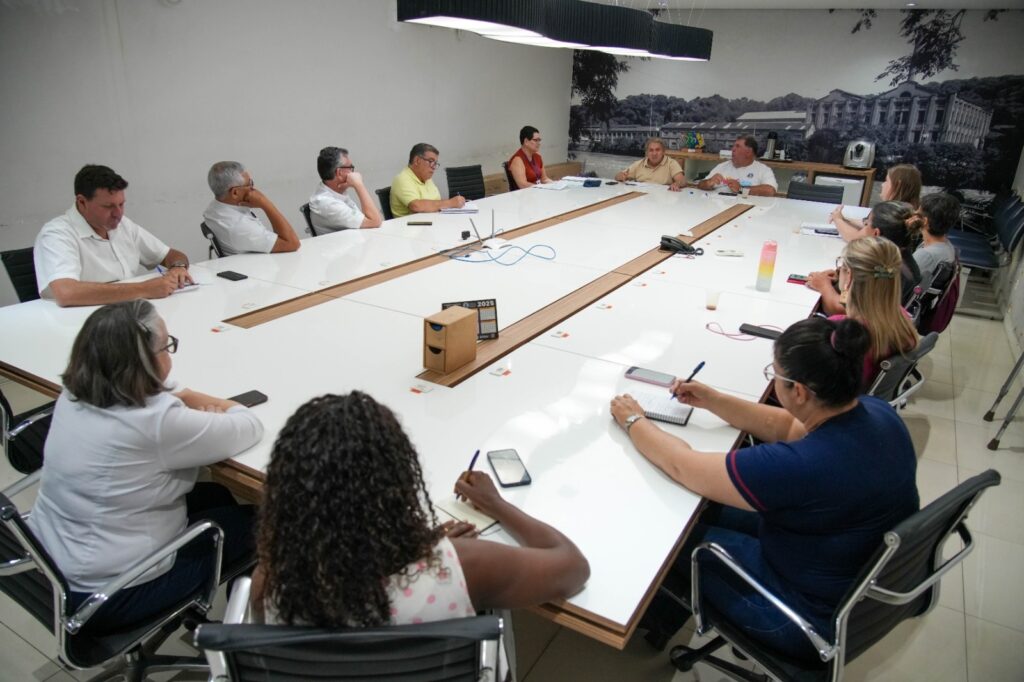 A cena retrata uma reunião em uma sala de conferências, com um grupo de pessoas sentadas ao redor de uma longa mesa branca. A iluminação é clara, vindo de um painel de luzes moderno no teto. À esquerda, um homem de camisa branca está escrevendo em um caderno, com um celular ao seu lado. Mais adiante, outro homem, também de camisa branca, apoia o queixo na mão, pensativo. Ao lado dele, um homem de camisa amarela observa algo à frente, com um caderno aberto. Do outro lado da mesa, em destaque, uma mulher de camisa branca com óculos está sentada, com as mãos apoiadas na mesa e um celular ao seu lado. Ao seu lado, uma mulher com cabelos cacheados e uma blusa estampada está escrevendo em um caderno, com um celular e uma caneta à sua frente. Mais adiante, uma mulher de blusa azul escura está escrevendo em um caderno, concentrada, com um celular ao lado. Outras pessoas estão sentadas ao redor da mesa, algumas em primeiro plano e outras mais ao fundo, participando da reunião. A parede ao fundo é decorada com um grande mural em preto e branco, que parece retratar uma paisagem urbana com edifícios antigos e árvores. A atmosfera geral sugere um ambiente de trabalho formal e produtivo.