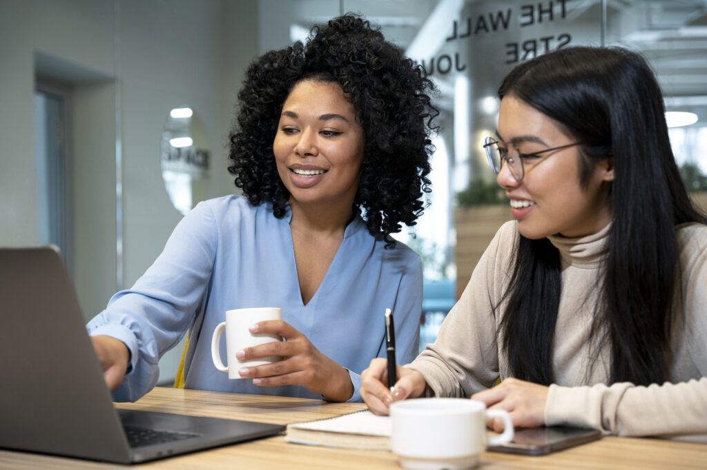 A imagem mostra duas mulheres sentadas a uma mesa, trabalhando juntas num portátil. A mulher à esquerda tem cabelo encaracolado e escuro e veste uma blusa azul clara. Ela está a segurar uma chávena branca numa mão e a usar a outra para interagir com o portátil. A mulher à direita tem cabelo preto comprido e liso e usa óculos. Ela está a usar um camisola bege de gola alta e a escrever num bloco de notas com uma caneta. Há outra chávena branca na mesa à sua frente. Ambas as mulheres parecem concentradas no seu trabalho, e há um ambiente de colaboração no escritório. O reflexo no vidro atrás delas sugere que estão num espaço de escritório moderno.