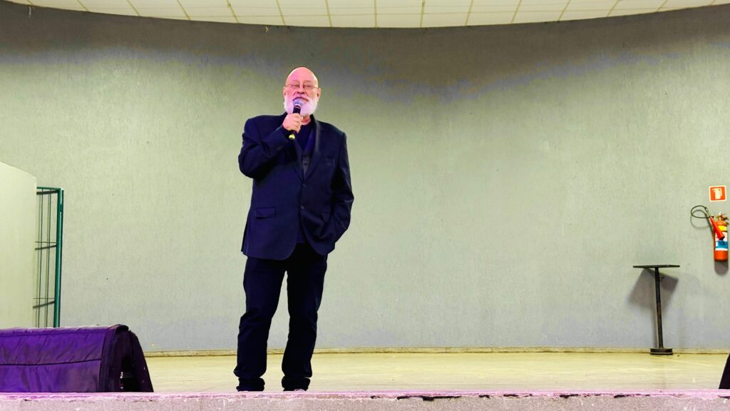 O homem, careca e com barba branca, está em um palco, segurando um microfone em sua mão direita e com a mão esquerda no bolso de seu terno escuro. Ele usa um terno azul escuro sobre uma camisa preta e calças pretas. O palco é iluminado e o fundo é uma parede cinza claro com uma linha horizontal escura no topo. À direita, há um extintor de incêndio vermelho pendurado na parede. À esquerda, há um objeto escuro coberto com um tecido roxo.