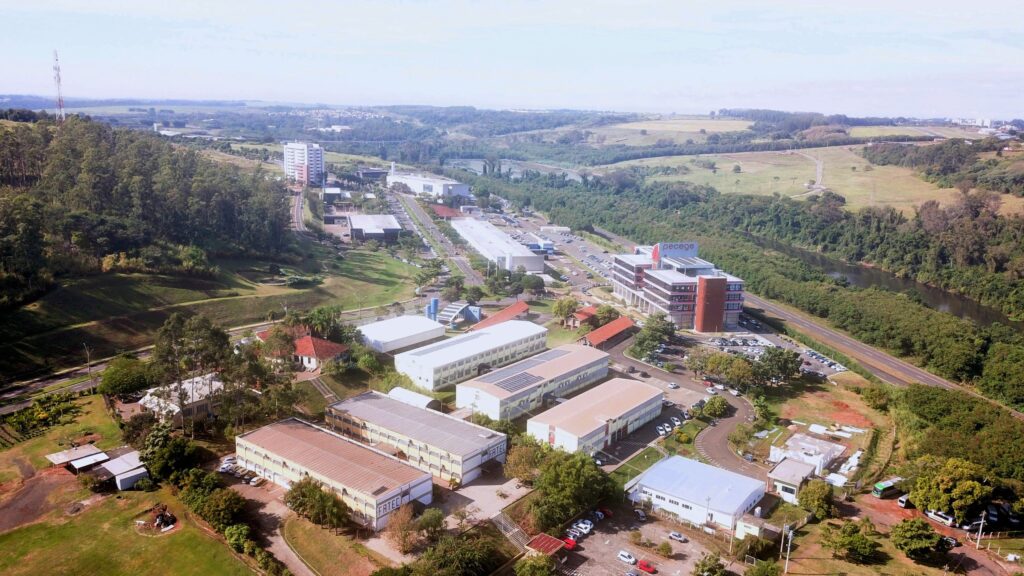 Na imagem aparece uma vista aérea com vários prédios espalhados por uma grande área rodeada por vegetação. No destaque aparece um prédio escrito PECEGE, em vermelho. Abaixo aparecem dois prédios escritos FATEC. No estacionamento em baixo de árvores aparecem alguns ônibus e carros estacionados.