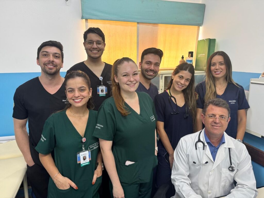 A imagem mostra um grupo de oito profissionais de saúde, todos sorrindo para a câmera. Eles parecem estar em um ambiente hospitalar ou clínico, com paredes claras e uma janela ao fundo. No primeiro plano, à direita, está um homem mais velho vestindo um jaleco branco e um estetoscópio pendurado no pescoço. Ele tem óculos e um sorriso amigável. Atrás dele e em pé, há sete pessoas mais jovens, a maioria vestindo uniformes verdes ou azuis escuros, típicos de profissionais de saúde. Há uma mistura de homens e mulheres no grupo. Alguns dos uniformes têm crachás com nomes e identificações. A iluminação é clara, vindo de cima e da janela ao fundo, criando um ambiente acolhedor.