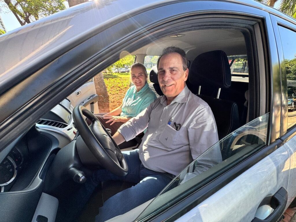 Em um dia ensolarado, um casal sorri para a câmera dentro de um carro. O homem, sentado no banco do motorista, veste uma camisa xadrez cinza e jeans. Ele tem uma caneta no bolso esquerdo da camisa. A mulher, sentada no banco do passageiro, usa uma camisa verde-água clara e jeans. Ambos parecem felizes e relaxados, com o sol iluminando seus rostos e o interior do veículo. A imagem foi tirada de fora do carro, com a porta do motorista aberta, permitindo uma visão clara dos ocupantes e do painel do carro.