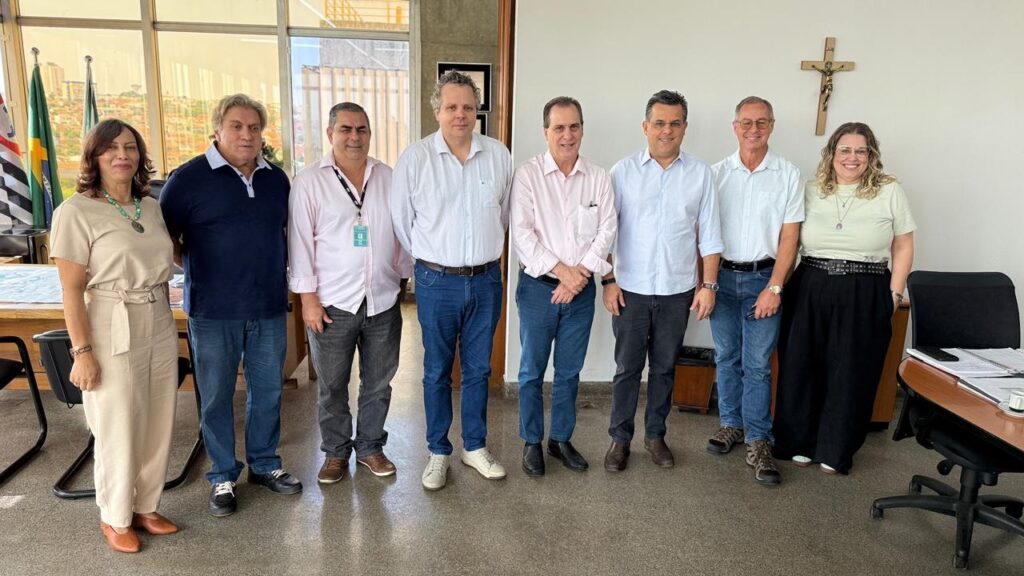 A imagem mostra um grupo de oito pessoas, cinco homens e duas mulheres, em pé em uma sala. Eles parecem estar posando para uma foto oficial. A mulher mais à esquerda veste um macacão bege com um cinto e um colar com um pingente grande. Ao lado dela, um homem com cabelo loiro e ondulado usa uma camisa polo azul marinho e calça jeans. O homem ao lado dele usa uma camisa social rosa clara e calça jeans escura. No centro da imagem, há mais três homens. O primeiro usa uma camisa social branca e calça jeans. O segundo, com cabelo escuro e óculos, veste uma camisa social rosa clara e calça jeans. O terceiro homem, também com óculos, usa uma camisa social azul clara e calça jeans. A mulher mais à direita está vestindo uma blusa verde clara, uma saia longa preta e óculos. Atrás do grupo, há uma janela grande com vista para uma cidade ao longe, e uma bandeira do estado de São Paulo e uma bandeira do Brasil estão visíveis. Há também uma cruz de madeira na parede. A sala parece ser um escritório, com mesas e cadeiras visíveis.
