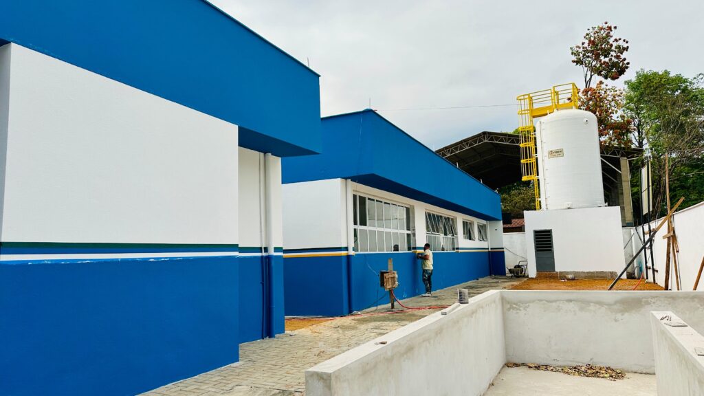 A imagem mostra a fachada de um edifício moderno, pintado em tons vibrantes de azul e branco. O edifício possui um design retilíneo com linhas arquitetônicas marcantes. Uma faixa azul escura percorre a base da construção, contrastando com as paredes brancas superiores. Uma linha fina amarela e outra verde delimitam a junção entre o azul e o branco, adicionando um toque de cor. No lado direito da imagem, destaca-se um grande tanque cilíndrico branco, com uma estrutura metálica amarela de acesso e uma plataforma no topo. Ao lado do tanque, há uma pequena edificação branca com uma porta cinza e uma grade de ventilação. Um trabalhador, vestindo uma camiseta amarela e calças escuras, está encostado na parede azul do edifício, parecendo estar em meio a alguma atividade de manutenção ou pintura. O chão em frente ao edifício é pavimentado com blocos de concreto intertravados.