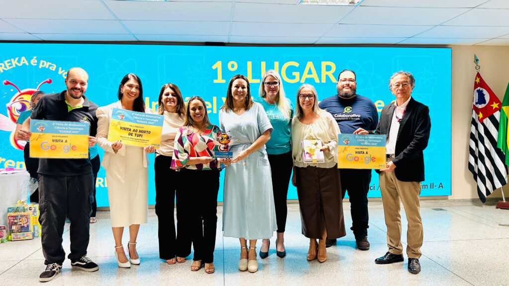 A imagem mostra um grupo de nove pessoas em pé, posicionado em frente a um grande painel azul com o texto "EUREKA é pra vo" e "1º LUGAR" em destaque. Várias pessoas no grupo estão segurando placas amarelas com a inscrição "VALE PRESENTE VISITA A Google" e "VALE PRESENTE VISITA AO HORTO DE TUPI", além de um troféu com a figura de uma abelha. As bandeiras do estado de São Paulo e do Brasil podem ser vistas à direita. O evento parece ser uma premiação para a "2ª Feira de Matemática, Ciência e Tecnologia 'EUREKA 2025'". As pessoas vestem roupas casuais e formais, e algumas sorriem para a câmera.