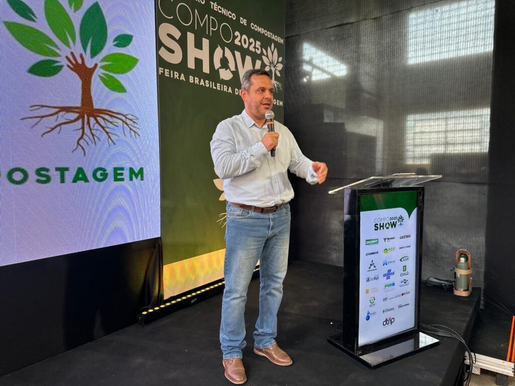A imagem mostra um homem em pé em um palco, falando em um microfone. Ele está vestindo uma camisa de botões de manga comprida azul clara e jeans. Atrás dele, há um grande banner com o texto "COMPOSTAGEM" e um logotipo de árvore estilizada. À sua direita, há um púlpito com vários logotipos de patrocinadores e o texto "COMPO 2025 SHOW". O palco está iluminado e há uma luz forte no fundo. A atmosfera geral sugere um evento ou conferência relacionada à compostagem.