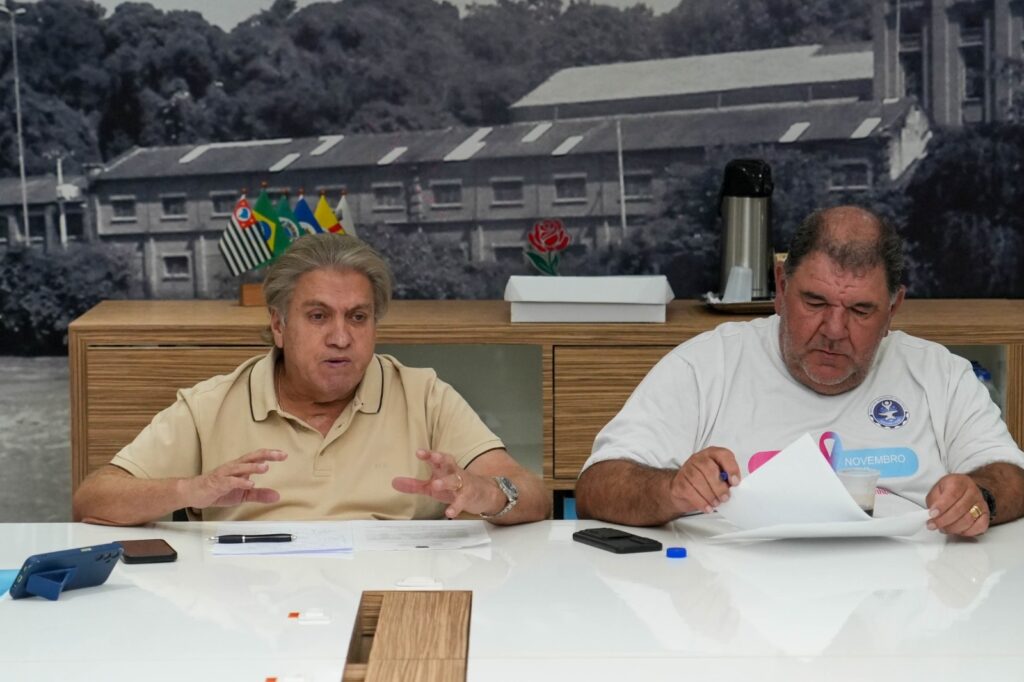 A imagem mostra dois homens sentados em uma mesa branca, com uma decoração de parede em preto e branco ao fundo que retrata um edifício industrial e árvores. O homem à esquerda, com cabelos grisalhos penteados para trás e vestindo uma camisa polo bege, gesticula com as mãos enquanto fala. Ele usa um relógio no pulso esquerdo. Na mesa à sua frente, há papéis, uma caneta preta, um celular em um suporte azul e um pequeno objeto retangular escuro. Atrás dele, em um móvel de madeira, há um conjunto de bandeiras em um suporte, incluindo a bandeira do estado de São Paulo e a bandeira do Brasil. O homem à direita, com cabelos escuros e calvície, veste uma camiseta branca com um selo circular azul e a palavra "NOVEMBRO" em letras grandes com uma fita rosa acima. Ele está curvado sobre alguns papéis que está segurando e olhando atentamente. Há um celular preto e uma tampa azul de garrafa na mesa à sua frente. Ao fundo, em um móvel de madeira, há uma garrafa térmica prateada e uma caixa branca.