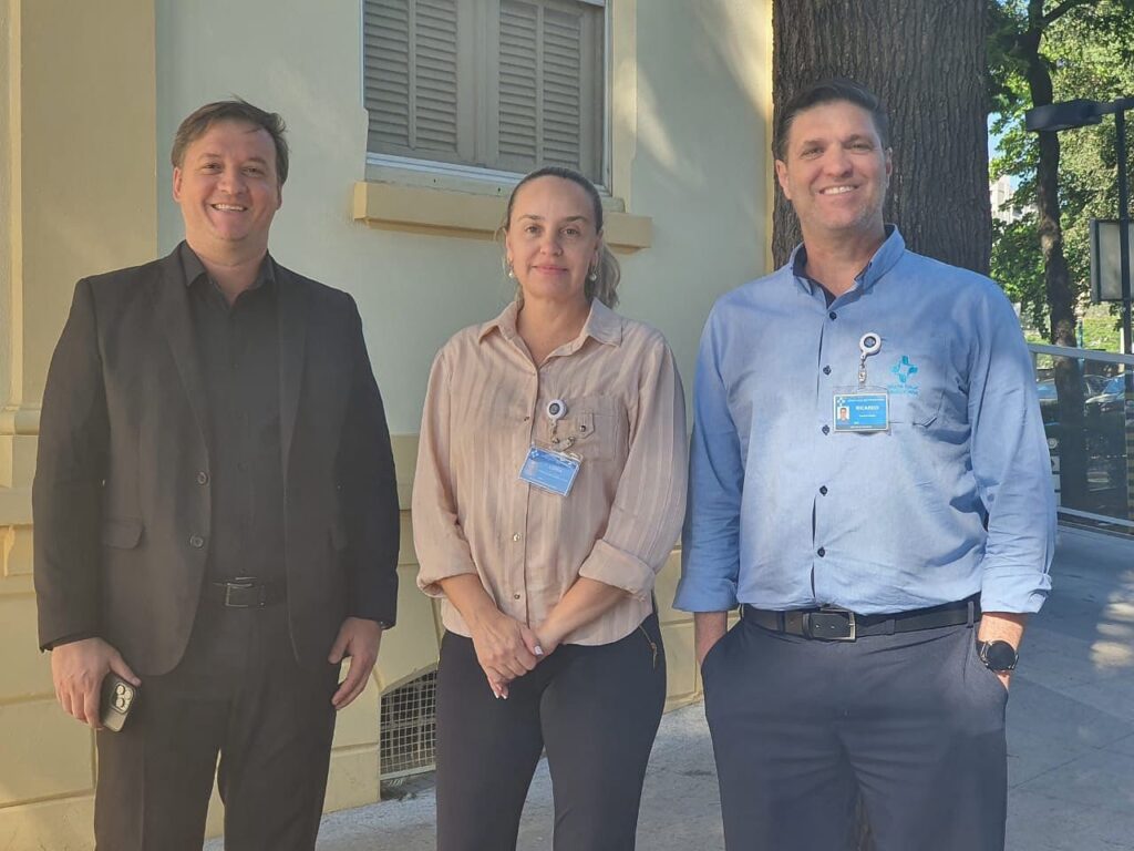 A imagem mostra três pessoas em pé, com um fundo de um prédio claro e uma árvore grande à direita. À esquerda, um homem veste um terno preto sobre uma camisa preta. Ele segura um celular na mão esquerda e sorri para a câmera. No centro, uma mulher veste uma camisa bege com listras finas e calças escuras. Ela tem um crachá preso à sua camisa e sorri levemente. À direita, um homem veste uma camisa azul clara com um emblema no peito e calças escuras. Ele também tem um crachá e sorri para a câmera, com as mãos nos bolsos. A luz do sol ilumina a cena, criando sombras e destacando os detalhes.