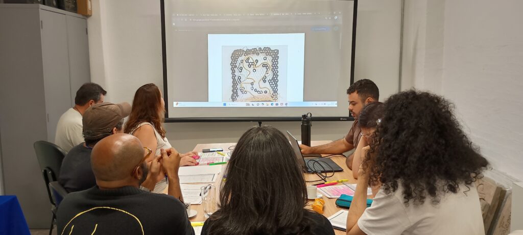 A imagem mostra um grupo de pessoas reunidas em torno de uma mesa, assistindo a uma apresentação em um projetor. A sala parece ser um espaço de trabalho ou sala de aula, com um quadro branco e armários ao fundo. No projetor, uma imagem de uma obra de arte abstrata está em exibição. A arte apresenta padrões intrincados e formas geométricas, com tons de cinza, bege e dourado. As pessoas na sala parecem engajadas na apresentação, com alguns olhando para o projetor, outros para seus laptops e cadernos. A iluminação na sala é artificial, vindo principalmente do projetor e de algumas luzes superiores. A atmosfera geral é de colaboração e aprendizado, com os participantes focados no conteúdo que está sendo apresentado. As pessoas na imagem têm diversas aparências, sugerindo um grupo heterogêneo.
