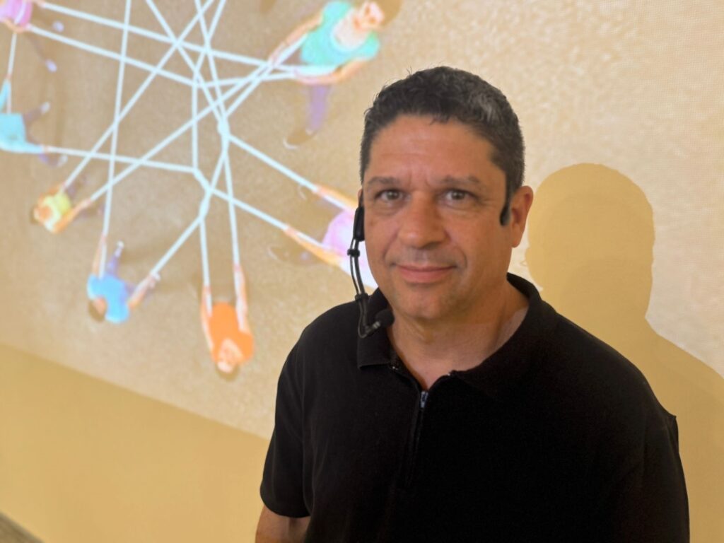 O homem na imagem veste uma camisa preta e tem um microfone preso à gola. Ele está de pé em frente a uma projeção de um grupo de pessoas dançando ou se movendo em um padrão circular, conectadas por linhas brancas. A projeção ilumina a parede atrás dele, criando uma sombra em forma de silhueta no lado direito da imagem. O homem olha diretamente para a câmera com uma expressão neutra.