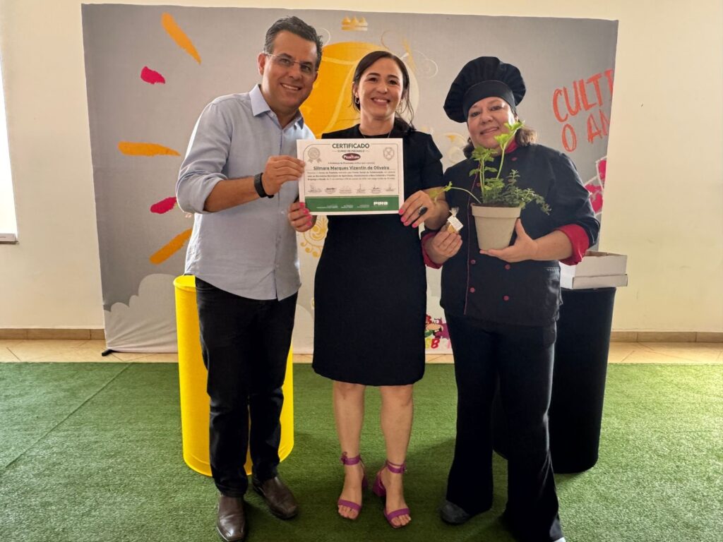Na imagem, três pessoas posam para uma foto, segurando um certificado e um vaso de plantas. À esquerda, um homem sorri para a câmera, vestindo uma camisa azul clara e calças pretas. Ele segura um certificado com as duas mãos. No centro, uma mulher de vestido preto e sandálias roxas também sorri. Ela segura o certificado com a mão esquerda, enquanto a mão direita repousa sobre a mão do homem. À direita, uma mulher com um chapéu de chef e um uniforme de chef preto com detalhes vermelhos segura um vaso de plantas com a mão direita. Ela sorri levemente para a câmera. Ao fundo, um painel com desenhos coloridos e um texto parcialmente visível que diz "CULT O A". O chão é coberto por um gramado artificial verde.