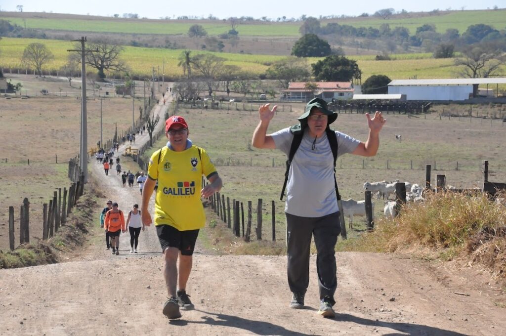 A imagem mostra um grupo de pessoas caminhando em uma estrada de terra rural. Dois homens estão em primeiro plano, um vestindo uma camiseta amarela e boné vermelho, e o outro com um chapéu verde. Ambos parecem estar caminhando em direção ao espectador. Atrás deles, uma longa fila de pessoas caminha pela estrada que se estende por uma paisagem rural com campos verdes e algumas construções ao longe. Há postes de eletricidade ao longo da estrada e cercas de madeira nas laterais. Ao fundo, um rebanho de gado branco pode ser visto em um pasto. O céu está claro e o dia parece ensolarado.