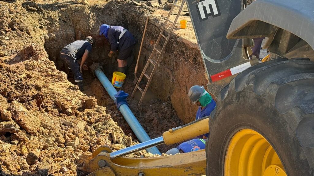 A imagem mostra uma equipe de trabalhadores em uma vala aberta, realizando trabalhos de infraestrutura. Vários homens estão agachados, trabalhando em um grande tubo azul que está parcialmente enterrado na terra. Uma retroescavadeira amarela está posicionada ao lado da vala, com sua pá frontal visível na parte inferior da imagem. Há uma escada de metal encostada na lateral da vala, sugerindo que os trabalhadores precisam subir e descer dela. Um balde amarelo com a marcação "GEN" está próximo ao tubo. O solo ao redor é de cor marrom-avermelhada e parece solto, com algumas pedras visíveis. Os trabalhadores estão vestidos com roupas de trabalho e alguns usam capacetes de segurança. A cena geral indica um trabalho de construção ou manutenção de tubulações em andamento.