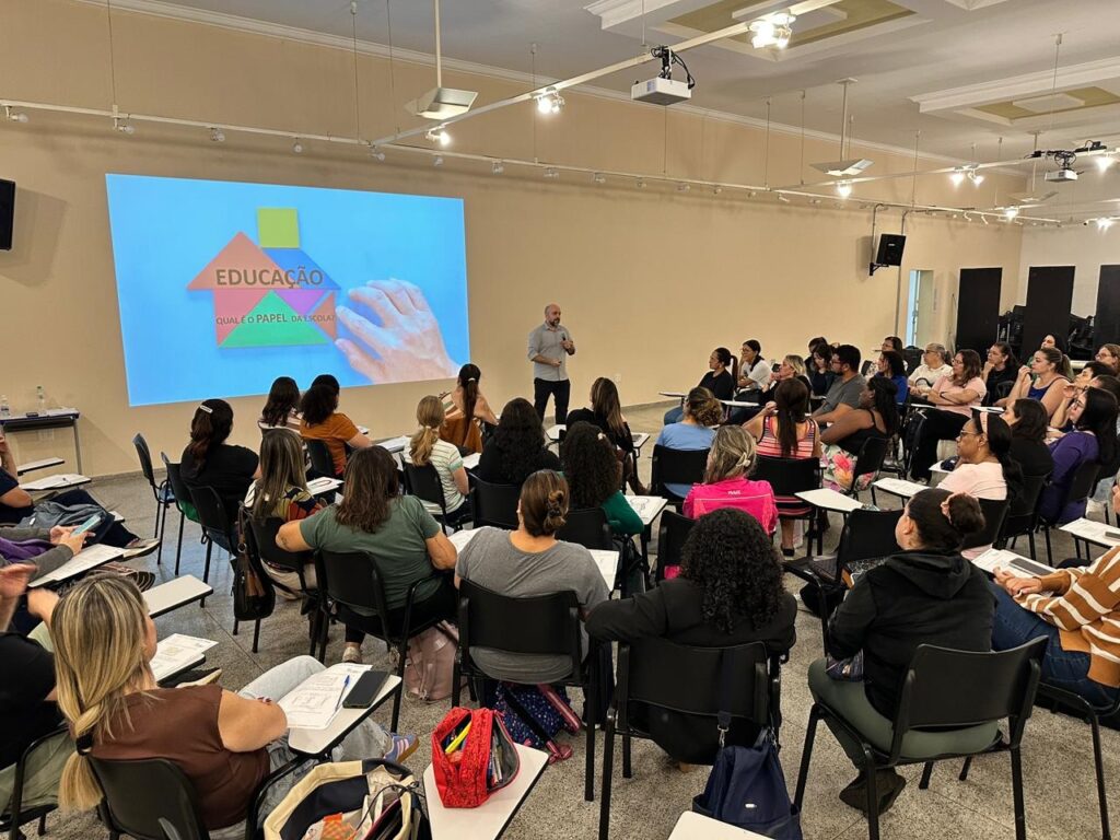 A palestra está sendo realizada em uma sala com muitas pessoas sentadas em cadeiras, olhando para uma tela grande. A tela exibe a palavra "EDUCAÇÃO" escrita em cores vibrantes, com um ícone de casa formado por blocos coloridos. Abaixo da palavra "EDUCAÇÃO", há uma pergunta escrita em português: "QUAL É O PAPEL DA ESCOLA?". Um homem está em pé perto da tela, provavelmente o palestrante, gesticulando enquanto fala. A audiência parece composta majoritariamente por mulheres, de diversas idades, e algumas estão anotando em cadernos ou olhando atentamente para o palestrante. O ambiente é iluminado por luzes no teto e a sala parece ser um local de treinamento ou conferência, com mesas e cadeiras dispostas em fileiras.