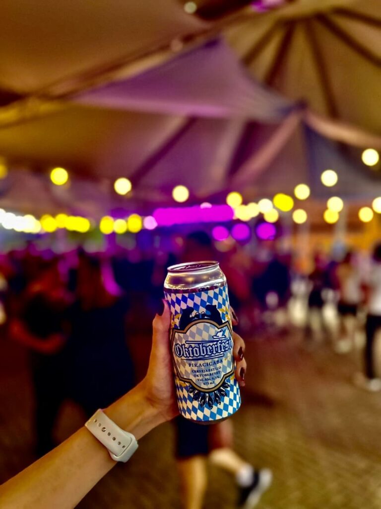A imagem mostra uma lata de cerveja "Oktoberfest" sendo segurada por uma mão com unhas pintadas de vermelho escuro. A lata tem um design xadrez azul e branco, com o logotipo da Oktoberfest em destaque. O fundo está desfocado, mas sugere um ambiente festivo com luzes coloridas (amarelas, roxas e rosas) e pessoas dançando ou se reunindo. Há também uma estrutura de teto decorativa com tecidos suspensos. A pessoa que segura a lata usa um smartwatch branco no pulso.