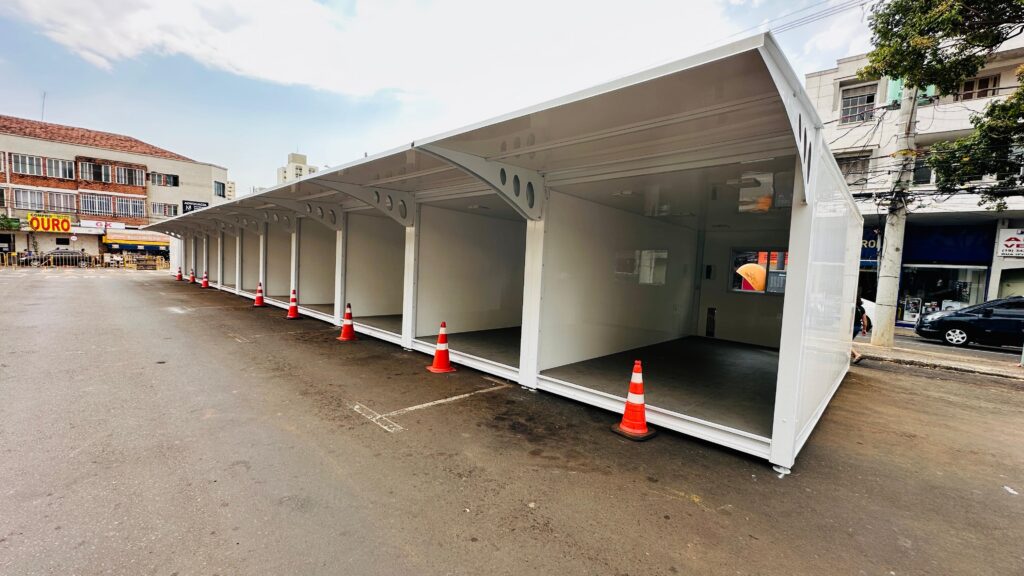 A imagem mostra uma estrutura modular branca, composta por várias baias ou cubículos interligados, que parece estar montada em uma área de asfalto, possivelmente uma rua ou estacionamento. A estrutura é longa e baixa, com um telhado inclinado que se estende sobre cada unidade. As baias são abertas na frente, revelando um interior branco e vazio. Vários cones de trânsito laranja e brancos estão posicionados no chão, em frente a cada baia, sugerindo que a área pode ser usada para algum tipo de serviço ou evento que requer organização de filas ou controle de acesso. Ao fundo, observam-se edifícios comerciais e residenciais, com algumas árvores e postes de iluminação pública. Uma rua com carros estacionados e em movimento é visível à direita da estrutura. O céu está parcialmente nublado.