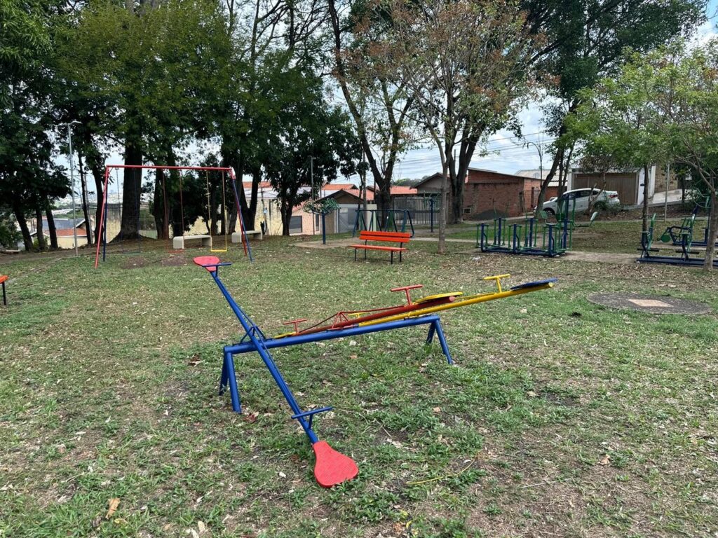 A imagem mostra um parquinho infantil em um dia nublado. No primeiro plano, há um gangorra azul com assentos amarelos e vermelhos. Ao fundo, vemos um balanço vermelho e amarelo, alguns bancos e outros equipamentos de playground. Há árvores ao redor do parquinho e algumas casas e carros ao fundo. A grama está um pouco alta e há algumas folhas caídas no chão.