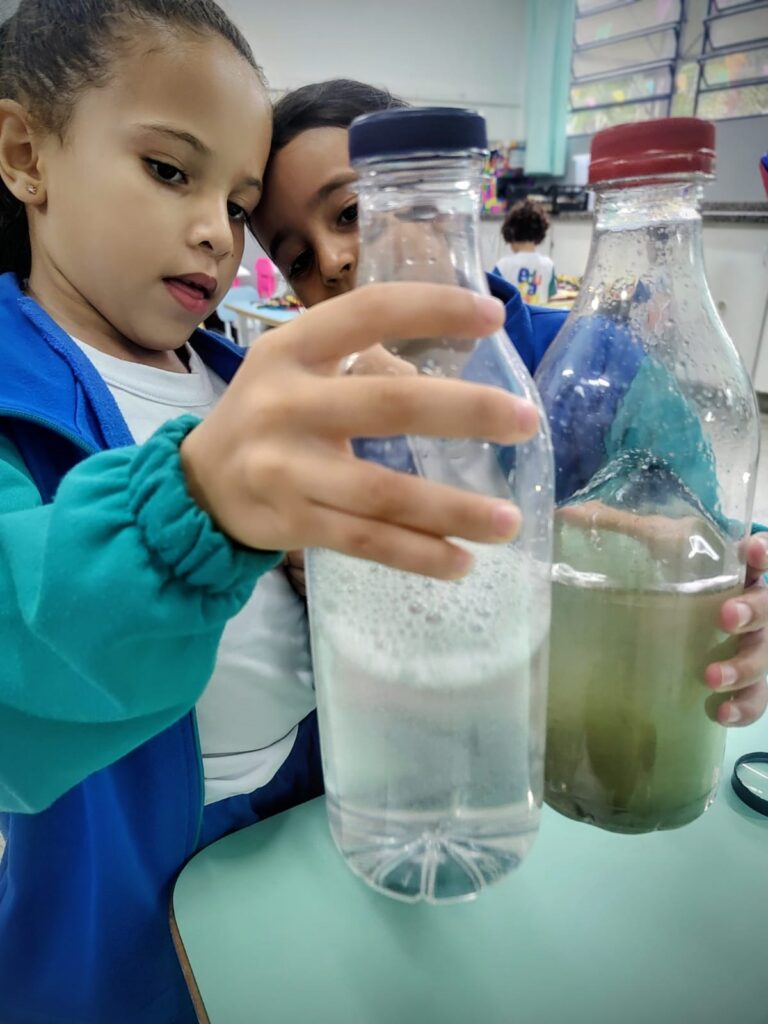 A imagem mostra duas crianças em uma sala de aula, provavelmente em uma atividade científica. Elas estão segurando duas garrafas plásticas transparentes com líquidos. A criança da esquerda, uma menina com cabelo escuro preso e usando uma jaqueta azul e branca, está olhando atentamente para a garrafa em suas mãos. Esta garrafa contém um líquido transparente com muitas bolhas na parte superior, sugerindo que algo foi misturado ou agitado. A criança da direita, um menino com cabelo escuro e usando uma jaqueta azul, está ao lado dela, também olhando para as garrafas. Ele segura uma segunda garrafa, que contém um líquido de cor verde-amarelada, com alguns sedimentos no fundo. A tampa desta garrafa é vermelha, enquanto a outra é azul. As crianças parecem estar engajadas em uma demonstração ou experimento, possivelmente relacionado à separação de substâncias, reações químicas ou filtragem. O ambiente sugere uma sala de aula, com mesas verdes e outras crianças e materiais visíveis ao fundo.