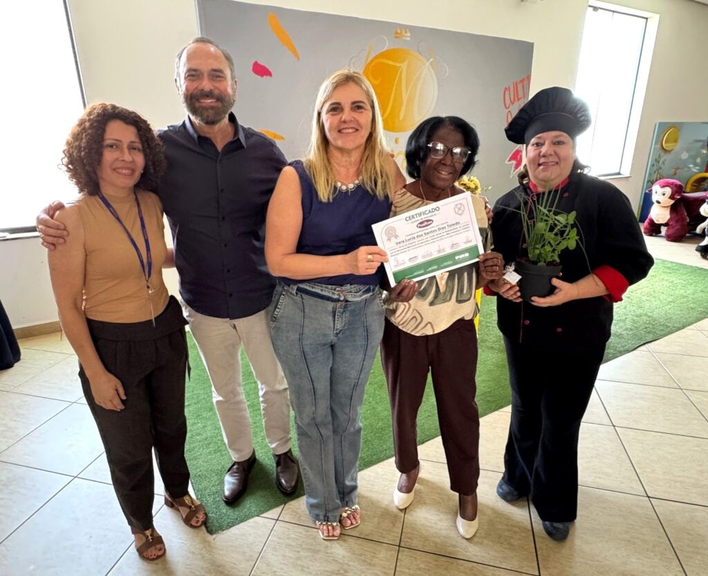 A imagem mostra cinco pessoas em um ambiente interno, possivelmente uma sala de eventos ou uma escola. No centro, um homem e duas mulheres estão abraçados, sorrindo para a câmera. O homem usa uma camisa azul escura e calça bege. A mulher à sua esquerda veste uma blusa bege e calça preta, com um crachá pendurado no pescoço. A mulher à sua direita usa uma blusa azul e calça jeans. As outras duas pessoas estão mais à direita. Uma delas, uma mulher negra com óculos, segura um certificado. A última pessoa, vestida com um uniforme de chef preto e vermelho, segura um pequeno vaso com plantas. O fundo da imagem apresenta uma parede decorada com desenhos coloridos e uma janela à direita. O chão é uma combinação de piso quadriculado e gramado artificial.