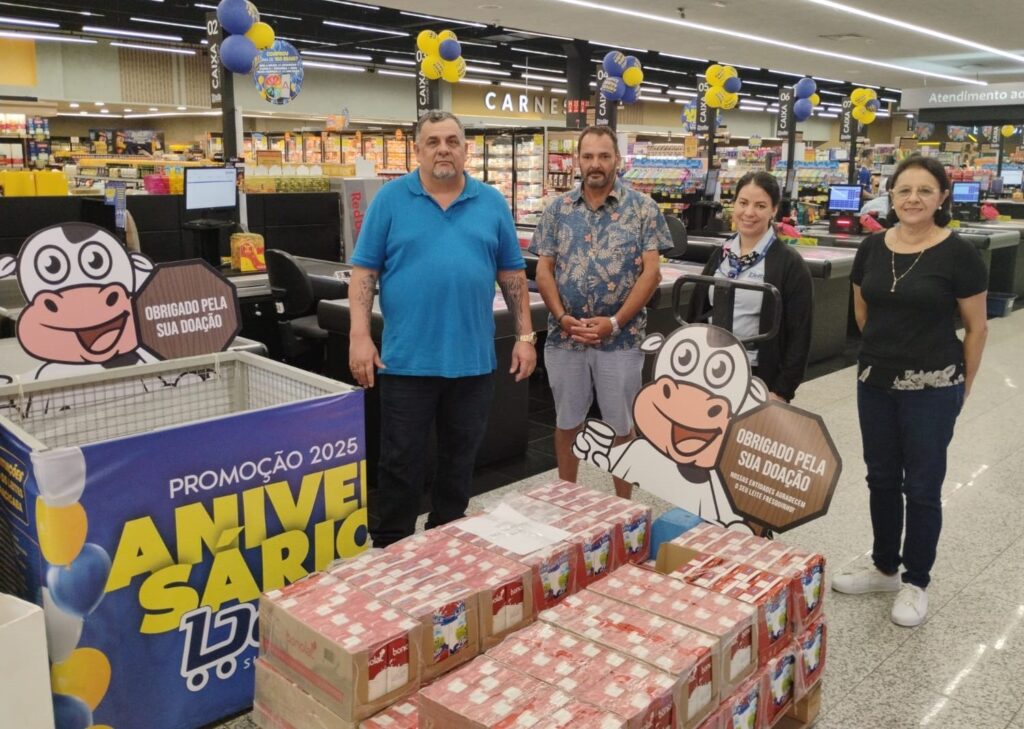 Em um supermercado, quatro pessoas estão em pé perto de caixas de leite e um ponto de coleta de doações. O ponto de coleta é um cesto com uma placa de animação de vaca ao lado, que diz "Obrigado pela sua doação". O cesto está decorado com um banner que diz "Promoção 2025 Aniversário" com balões azuis e amarelos. Uma grande quantidade de caixas de leite está empilhada em frente ao ponto de coleta. As pessoas parecem ser funcionários do supermercado, com uma delas usando um crachá. Ao fundo, estantes de produtos e caixas registradoras são visíveis.