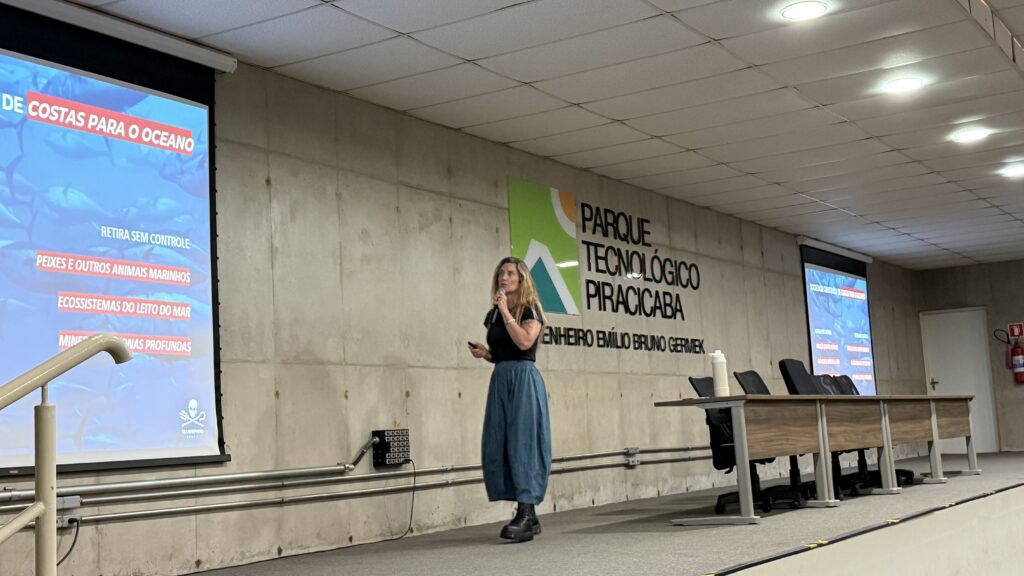 Uma mulher está em pé sobre um palco, segurando um microfone e fazendo uma apresentação. Atrás dela há uma parede de concreto com o nome “Parque Tecnológico Piracicaba Engenheiro Emílio Bruno Germek” e um logotipo colorido. No palco, há três cadeiras vazias e uma mesa com microfones, sugerindo um ambiente de debate ou conferência. Duas telas grandes exibem um slide com o título “De Costas para o Oceano” e quatro tópicos: retirada sem controle, peixes e outros animais marinhos, ecossistemas do litoral e do mar, e minerais das profundezas. O cenário indica uma palestra sobre conservação marinha e os impactos da ação humana nos oceanos.