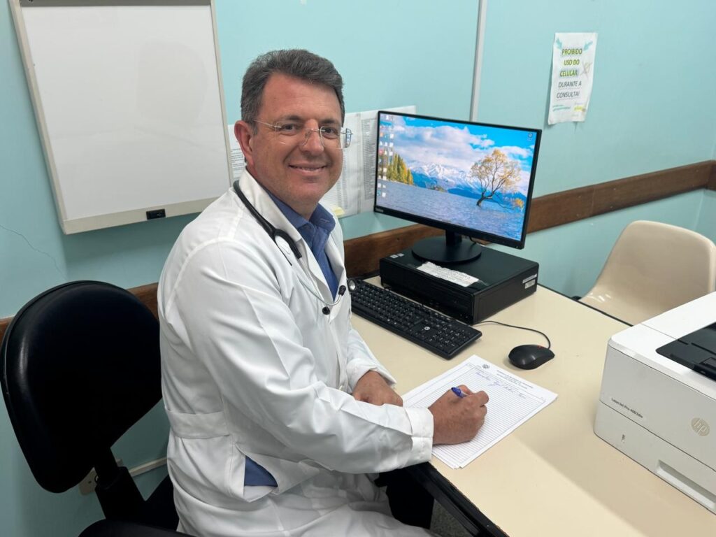 Um médico sorri para a câmera enquanto está sentado em sua mesa em um consultório médico. Ele está usando um jaleco branco sobre uma camisa azul e tem um estetoscópio pendurado em volta do pescoço. Ele está escrevendo em um pedaço de papel com uma caneta azul. A mesa está equipada com um computador com uma imagem de paisagem na tela, um teclado, um mouse e uma impressora. Há também um quadro branco na parede atrás dele e um aviso em português que diz "PROIBIDO USO DO CELULAR DURANTE A CONSULTA" (Proibido usar o celular durante a consulta).