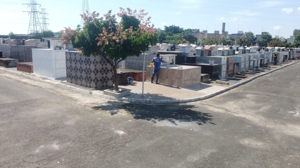 A imagem mostra um cemitério em um dia ensolarado. O céu está claro com algumas nuvens esparsas. Ao fundo, há torres de alta tensão e alguns edifícios. Em primeiro plano, um homem de camiseta azul e calça jeans está em frente a um jazigo, aparentemente limpando ou cuidando dele. Ele segura um balde amarelo em uma das mãos. Um pequeno arbusto com flores rosadas e folhas verdes está posicionado ao lado de um dos jazigos. O cemitério é composto por fileiras de jazigos de diferentes tamanhos e materiais, alguns com acabamento em azulejos, outros em mármore ou granito. A maioria dos jazigos tem um formato retangular, com alguns apresentando pequenas estruturas semelhantes a casinhas no topo. Um caminho de concreto contorna os jazigos, separando-os da rua asfaltada. As sombras das árvores e das estruturas projetam-se sobre o chão.