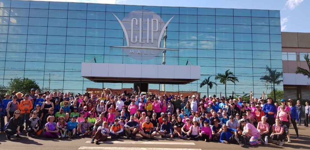 A imagem mostra um grande grupo de pessoas em frente a um prédio com fachada de vidro espelhado. O prédio tem uma grande placa com o nome "CLIP MALL" em letras estilizadas. As pessoas parecem estar reunidas para um evento, possivelmente uma caminhada ou corrida, pois muitas delas estão vestindo roupas esportivas e algumas têm mochilas. Há uma mistura de homens e mulheres de várias idades. O dia parece ensolarado, com céu azul e algumas nuvens. Palmiers e árvores são visíveis ao redor do prédio. A maioria das pessoas está olhando para a câmera e algumas estão acenando, indicando que a foto foi tirada de forma planejada. A multidão é densa, com pessoas agrupadas em diferentes níveis, algumas sentadas no chão e outras em pé.