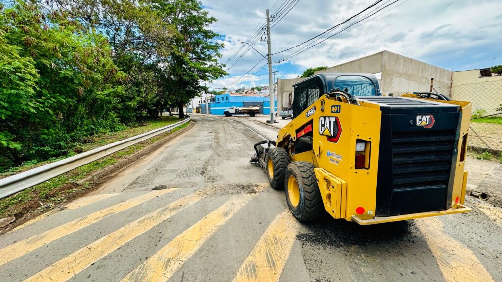 A imagem mostra uma máquina amarela da marca CAT, possivelmente uma retroescavadeira compacta, estacionada em uma rua de asfalto. A máquina está posicionada na lateral direita da imagem, com sua traseira voltada para o observador. A rua parece estar em processo de manutenção ou construção, com marcas de pneus e pedaços de asfalto espalhados. Ao fundo, a rua se estende em direção a um prédio azul e outras construções. Há árvores verdes exuberantes e uma cerca de metal ao longo da lateral esquerda da rua. O céu está nublado, mas com alguns raios de sol que iluminam a cena. A pintura amarela e preta da máquina CAT se destaca contra o ambiente ao redor.