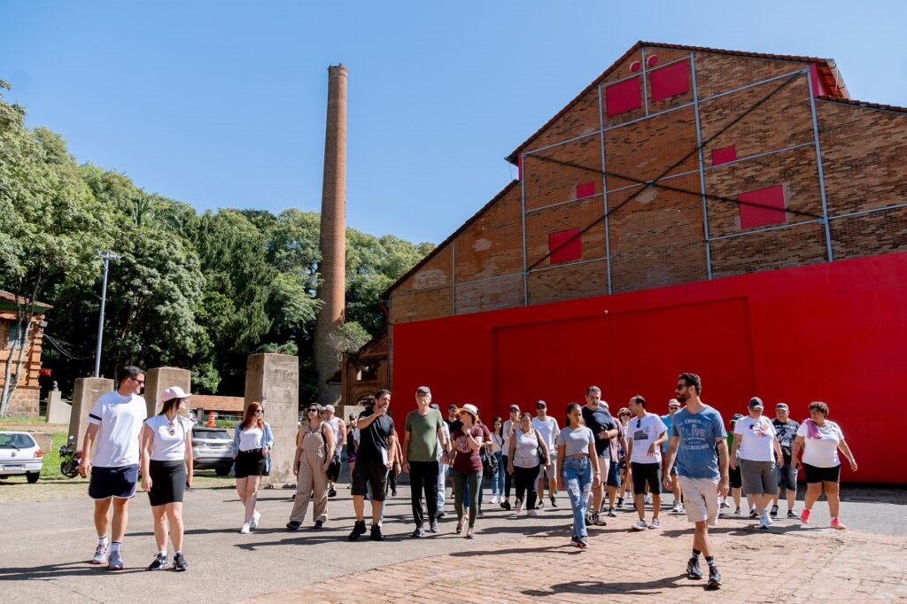 A imagem mostra um grupo de pessoas caminhando em um dia ensolarado, possivelmente em um tour guiado. Ao fundo, há um edifício de tijolos com uma grande seção vermelha vibrante e uma chaminé alta de tijolos. Árvores verdes exuberantes cercam a área, contrastando com o céu azul claro. As pessoas estão vestidas com roupas casuais de verão, e algumas parecem estar conversando entre si enquanto se movem. A cena sugere um ambiente de lazer ou cultural ao ar livre.