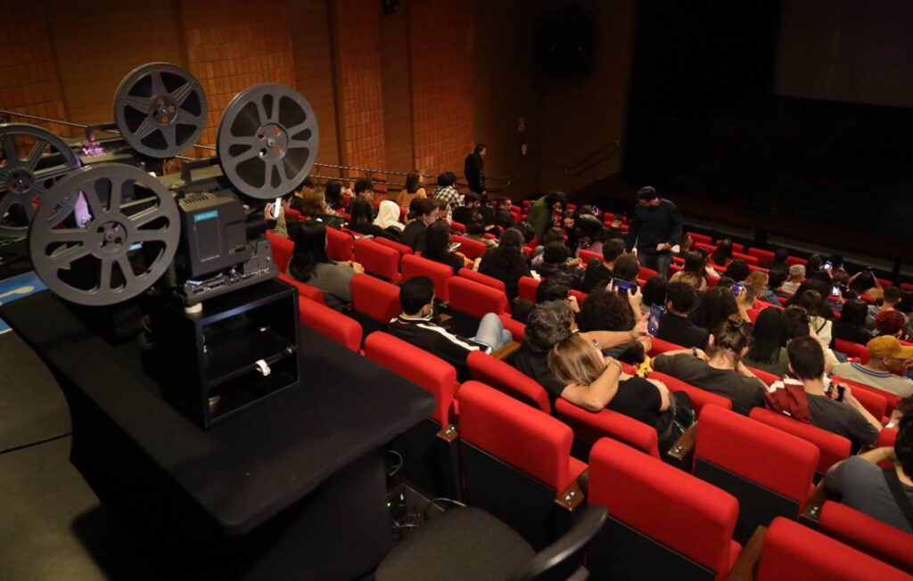 A imagem retrata uma sala de cinema com uma audiência sentada em poltronas vermelhas, aguardando o início de uma exibição. No primeiro plano, um projetor de filmes antigo, com seus rolos de filme característicos, está posicionado sobre uma mesa coberta por um pano preto. A iluminação é baixa, típica de um ambiente de cinema, com a tela principal ao fundo, escura e vazia. As pessoas na plateia estão em diferentes posições, algumas conversando, outras olhando para seus celulares, e algumas já acomodadas em suas poltronas. A atmosfera sugere a antecipação de um evento cinematográfico, com a presença do equipamento de projeção clássico adicionando um toque nostálgico à cena. A composição da imagem, com o projetor em destaque e a multidão ao fundo, cria um contraste entre o passado e o presente da experiência de assistir a filmes.