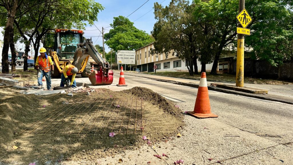 A imagem mostra uma cena de obras em uma rua, com dois trabalhadores usando coletes laranja e capacetes amarelos. Um trator retroescavadeira, da marca John Deere, está posicionado atrás deles. Há uma área escavada no chão, com uma malha de vergalhões de metal exposta, coberta parcialmente por terra e cascalho. Na lateral da rua, cones de trânsito laranja e brancos sinalizam a área de trabalho. Ao fundo, um edifício com uma placa branca que indica "Unidade Básica de Atendimento UBA - Piracicaba" e um número de telefone. Um poste de luz amarelo com sinalização de pedestres e a indicação "A 100 m" também está visível. O céu está claro e o dia parece ensolarado, com árvores frondosas ao redor.