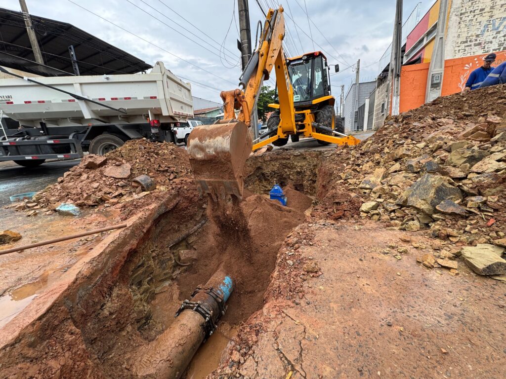 A imagem mostra uma cena de obra em uma rua, com uma escavadeira amarela em ação. * A escavadeira está removendo terra de uma vala profunda onde uma tubulação de água grande e suja está exposta. A tubulação parece ter emendas e conexões metálicas. * Ao lado da vala, há uma pilha de terra e pedras que foram escavadas. Uma grande caminhão basculante branco está estacionado próximo, provavelmente para transportar o material removido. * No fundo da vala, um hidrante azul está visível, indicando que a obra está relacionada ao abastecimento de água. * No lado direito da imagem, há uma construção com paredes coloridas e dois trabalhadores observando a obra. O céu está nublado, sugerindo um dia de trabalho em condições climáticas variadas.