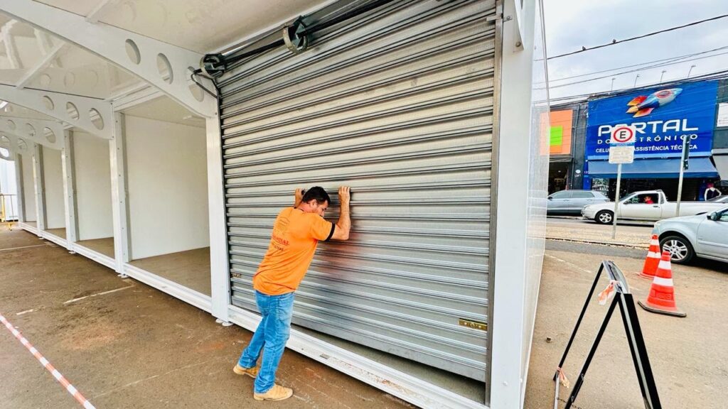 A imagem mostra um homem de camiseta laranja e calça jeans empurrando uma porta de metal em um local que parece ser uma loja ou um conjunto de quiosques. A porta de metal é do tipo "roly-poly" e está parcialmente fechada. Ao fundo, vê-se a fachada de uma loja com uma placa azul escrita "PORTAL ELETRÔNICO" e "CELULAR ASSISTÊNCIA TÉCNICA". Há carros estacionados na rua e cones de sinalização laranja e brancos. A estrutura onde o homem está parece ser modular, com várias divisórias internas visíveis.