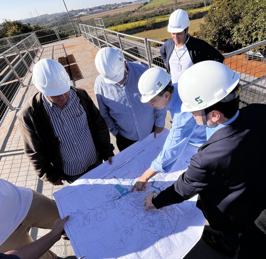 A imagem mostra um grupo de homens de capacete olhando um mapa. Eles parecem estar em um local elevado, com uma vista para uma paisagem urbana e rural ao fundo. A maioria dos homens está usando capacetes brancos com um logotipo verde. Um deles, de terno escuro, aponta para uma área específica do mapa, enquanto outro, de camisa azul clara, também indica algo com o dedo. Há uma cerca de metal ao redor da área onde eles estão. O mapa parece detalhar uma cidade, com ruas, rios e áreas construídas. O texto no mapa indica "OBRAS ESTRATÉGICAS" e "SEMAE / PREFEITURA MUNICIPAL DE PIRACICABA".