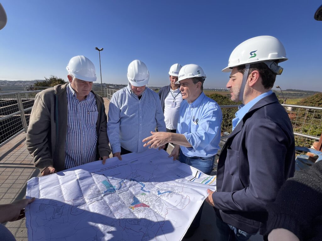 A imagem mostra um grupo de cinco homens, todos usando capacetes brancos, reunidos em torno de um mapa grande e detalhado. Eles estão em uma área externa com um céu azul claro e um pouco de vegetação ao fundo. O homem mais próximo à direita, vestindo um blazer azul escuro sobre uma camisa azul clara, está gesticulando com a mão direita sobre o mapa, parecendo explicar algo. Ele tem um capacete branco com o logotipo "S" verde. Os outros homens, vestindo camisas de botões em tons de azul e xadrez, também usam capacetes brancos, alguns com o mesmo logotipo "S". Um deles tem uma crachá pendurado no pescoço. O mapa parece ser um plano de obras estratégicas, com diferentes áreas coloridas (azul, vermelho e verde) e linhas que indicam rotas ou infraestrutura. A legenda do mapa menciona "OBRAS ESTRATÉGICAS" e "SEMAE / PREFEITURA MUNICIPAL DE PIRACICABA", sugerindo que se trata de um projeto de saneamento ou infraestrutura urbana na cidade de Piracicaba. A atmosfera parece ser de uma reunião de trabalho ou inspeção de obra, com os participantes focados na análise do mapa para discutir os detalhes do projeto.
