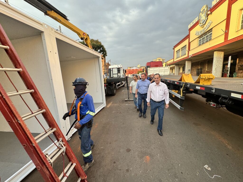No primeiro plano, à esquerda, há uma escada vermelha apoiada em uma estrutura branca, que parece ser um módulo pré-fabricado ou uma cabine. Um trabalhador vestindo um uniforme azul, capacete preto, máscara facial escura e cinto de segurança laranja está em pé ao lado da estrutura, com o olhar voltado para a direita. Ele usa luvas pretas e calças jeans com faixas refletivas amarelas nos tornozelos. Em segundo plano, um grupo de homens caminha pela rua. O homem mais próximo à câmera, à direita do grupo, veste uma camisa social clara e calças jeans escuras. Ele caminha em direção à câmera. Atrás dele, outros homens caminham em fila, alguns vestindo camisas sociais e outros roupas mais casuais. Ao fundo, há vários caminhões, incluindo um caminhão com uma plataforma de transporte e um caminhão com uma caçamba vermelha. À direita da imagem, um edifício amarelo com detalhes em vermelho e branco se destaca, com a inscrição "MERCADO ARAÇATUBA" em letras grandes. A arquitetura do edifício sugere um local comercial ou de serviços. A cena geral indica uma operação de transporte, instalação ou manutenção de uma estrutura modular, com a presença de trabalhadores e, possivelmente, autoridades ou interessados observando o processo.