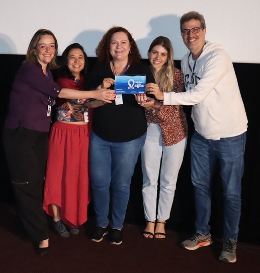 A imagem mostra cinco pessoas em pé, sorrindo e posando juntas. Elas parecem estar em um evento ou apresentação, com uma tela branca ao fundo. Da esquerda para a direita: * A primeira pessoa é uma mulher com cabelo loiro, vestindo uma blusa roxa e calças pretas. * A segunda pessoa é uma mulher com cabelo escuro, usando uma blusa estampada e uma saia longa vermelha. * A terceira pessoa é uma mulher com cabelo cacheado e ruivo, vestindo uma blusa preta e calças jeans. Ela está segurando um pequeno certificado ou placa azul com um desenho de onda e texto em branco. * A quarta pessoa é uma mulher com cabelo loiro, usando uma blusa estampada com manchas e calças jeans claras. * A quinta pessoa é um homem com cabelo escuro e óculos, vestindo um moletom branco com capuz e calças jeans.