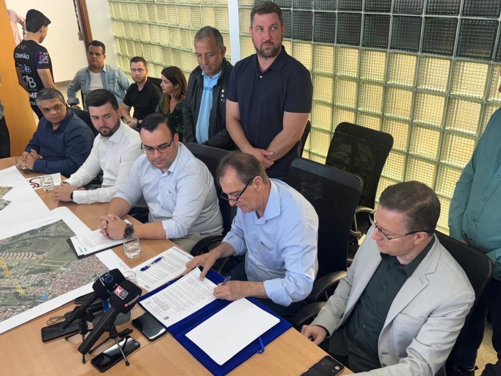 A imagem mostra um grupo de pessoas reunidas em torno de uma mesa em uma sala de reuniões. No centro da mesa, um homem de camisa azul claro está assinando um documento, enquanto outro homem de camisa clara e óculos o observa. Há também microfones de TV sobre a mesa, sugerindo que o evento pode ser uma coletiva de imprensa ou uma cerimônia de assinatura. Ao redor da mesa, outras pessoas estão sentadas ou em pé, observando a cena. Um mapa ou plano está parcialmente visível na mesa, indicando que o assunto da reunião pode estar relacionado a um projeto de desenvolvimento urbano ou de infraestrutura. O ambiente parece profissional, com paredes de vidro quadriculado ao fundo.