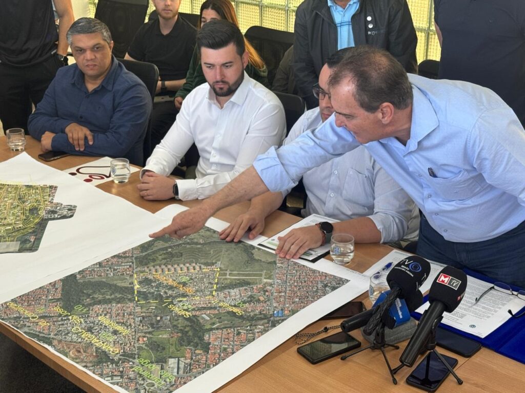 A imagem mostra um grupo de homens reunidos em torno de uma mesa com mapas e documentos. Um homem, vestindo uma camisa azul clara, está apontando para um dos mapas, explicando algo aos outros. Há também microfones de imprensa na mesa, sugerindo que o evento pode ser uma coletiva de imprensa ou uma reunião com cobertura midiática. As pessoas parecem estar engajadas na discussão, com expressões de concentração em seus rostos. Os mapas detalhados indicam que a reunião provavelmente trata de planejamento urbano, desenvolvimento de projetos ou alguma questão geográfica específica.