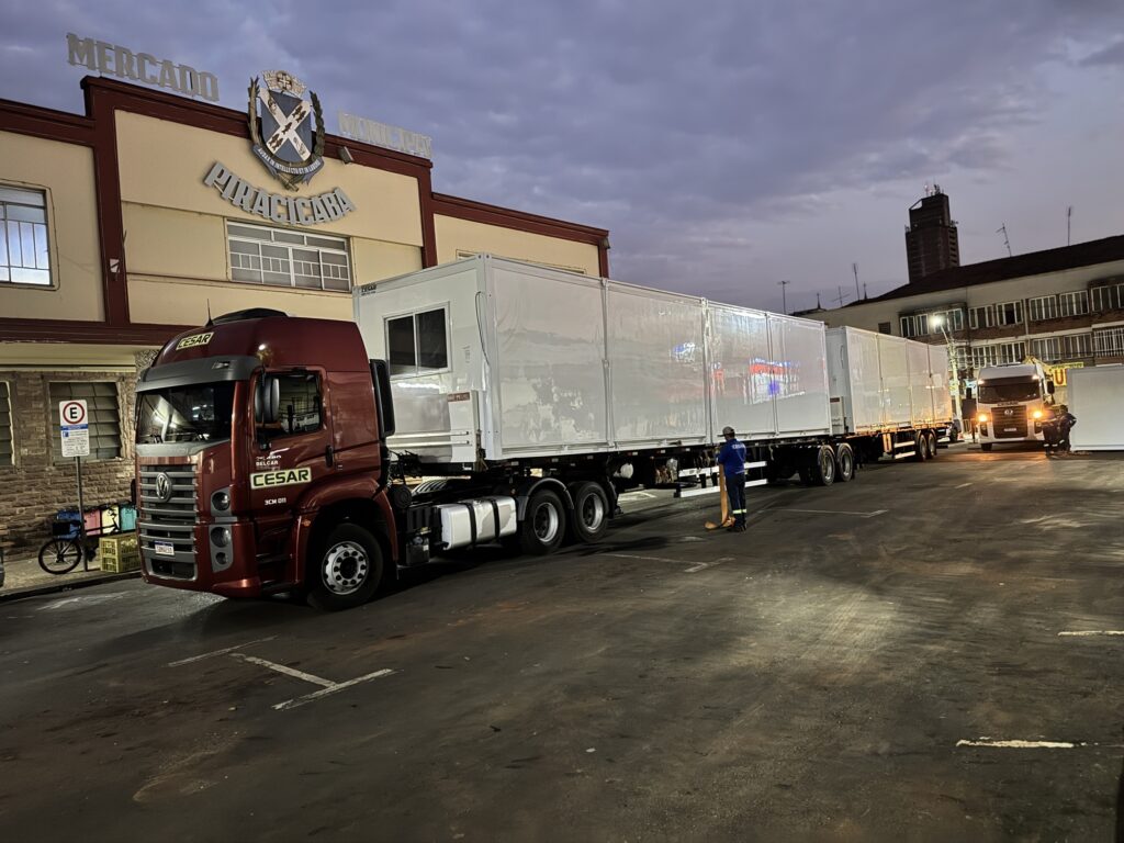A imagem mostra um estacionamento com vários caminhões. Um caminhão vermelho com o nome "CESAR" escrito na lateral está estacionado em primeiro plano, com seus trailers brancos estendendo-se para trás. Um homem em uniforme azul está parado ao lado dos trailers, segurando uma vassoura. Ao fundo, há um edifício com a palavra "MERCADO" escrita em letras grandes no topo e "PIRACICABA" abaixo. Outro caminhão branco está estacionado mais adiante no estacionamento, com suas luzes acesas. O céu está nublado, sugerindo que a foto foi tirada ao entardecer ou ao amanhecer. O estacionamento parece estar molhado, com reflexos das luzes dos caminhões e do edifício no chão.
