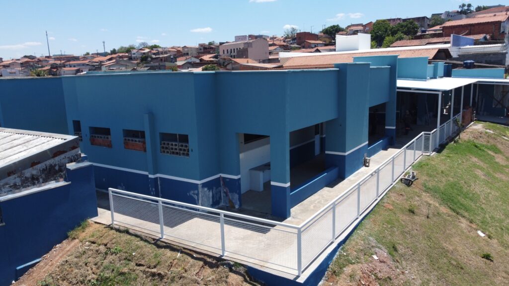 A imagem mostra um edifício azul com detalhes em branco, com uma estrutura em dois níveis. O nível inferior é pintado de azul escuro, enquanto o nível superior é azul claro. Há uma varanda coberta com um telhado branco que se estende ao longo do lado do edifício, com uma cerca branca que a delimita. O edifício está localizado em uma área montanhosa, com grama e terra visíveis no primeiro plano e ao redor da estrutura. Ao fundo, há uma coleção de casas com telhados vermelhos e marrons, sugerindo um bairro residencial. O céu está limpo e azul, com algumas nuvens dispersas. A arquitetura do edifício parece moderna, com linhas retas e formas geométricas. As janelas são retangulares e algumas delas parecem ser de blocos de concreto vazados. Há também uma entrada aberta no nível superior, que leva à varanda coberta.