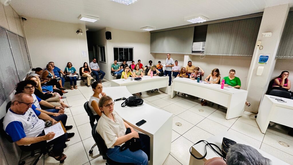 A imagem mostra uma sala de reunião com várias pessoas sentadas em cadeiras ao redor de mesas brancas. Um homem está de pé em frente a um grupo, aparentemente falando ou apresentando algo. Há um ar de formalidade, mas também de interação, com algumas pessoas observando atentamente o orador e outras conversando entre si. A iluminação é artificial, vinda de luminárias no teto, e as paredes são claras. O ambiente parece ser um escritório ou sala de conferências.