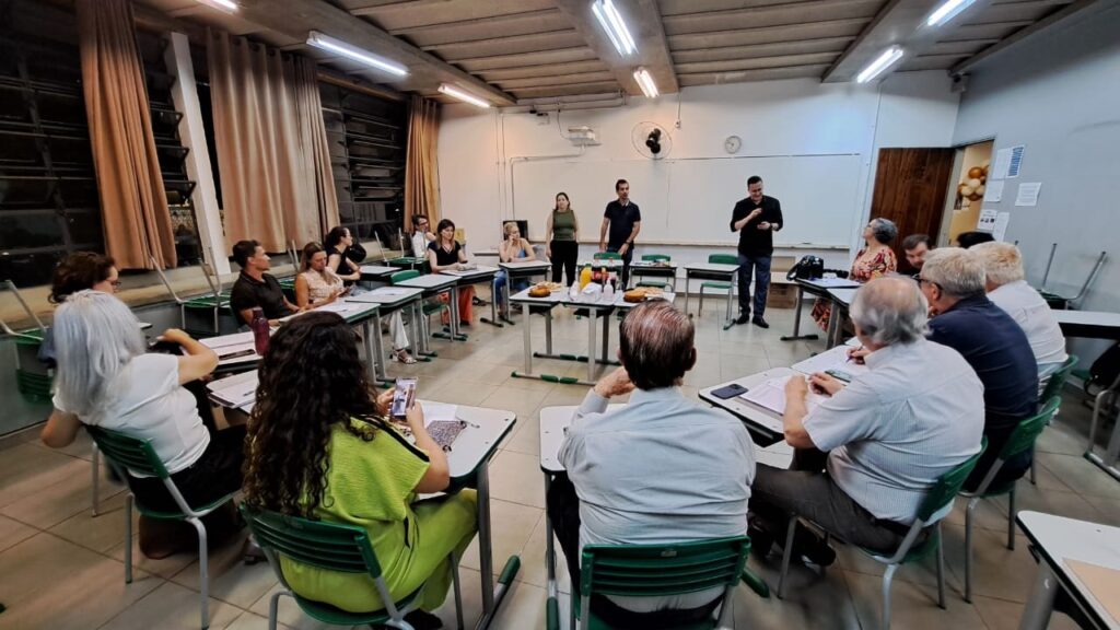 A imagem mostra um grupo de pessoas reunidas em uma sala de aula ou auditório. Há mesas dispostas em formato de U, com cadeiras ao redor. Algumas pessoas estão sentadas às mesas, enquanto outras estão em pé, em frente ao grupo. Há um quadro branco na parede atrás das pessoas em pé. Na mesa central, há alguns alimentos e bebidas, sugerindo um lanche ou confraternização. A iluminação é artificial, vinda de luminárias no teto. A atmosfera parece ser de um encontro formal, possivelmente uma reunião, palestra ou workshop.