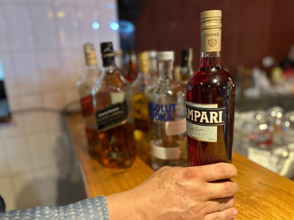 A imagem mostra uma mão segurando uma garrafa de Campari em primeiro plano. A garrafa é de vidro escuro e contém um líquido vermelho vibrante. A etiqueta do Campari é proeminente, com o nome da marca em letras grandes e douradas. Ao fundo, desfocadas, há várias outras garrafas de bebidas alcoólicas, incluindo uma garrafa de Johnnie Walker e uma garrafa de vodka Absolut. As garrafas estão dispostas em uma prateleira ou balcão de madeira. A iluminação sugere um ambiente de bar ou restaurante.