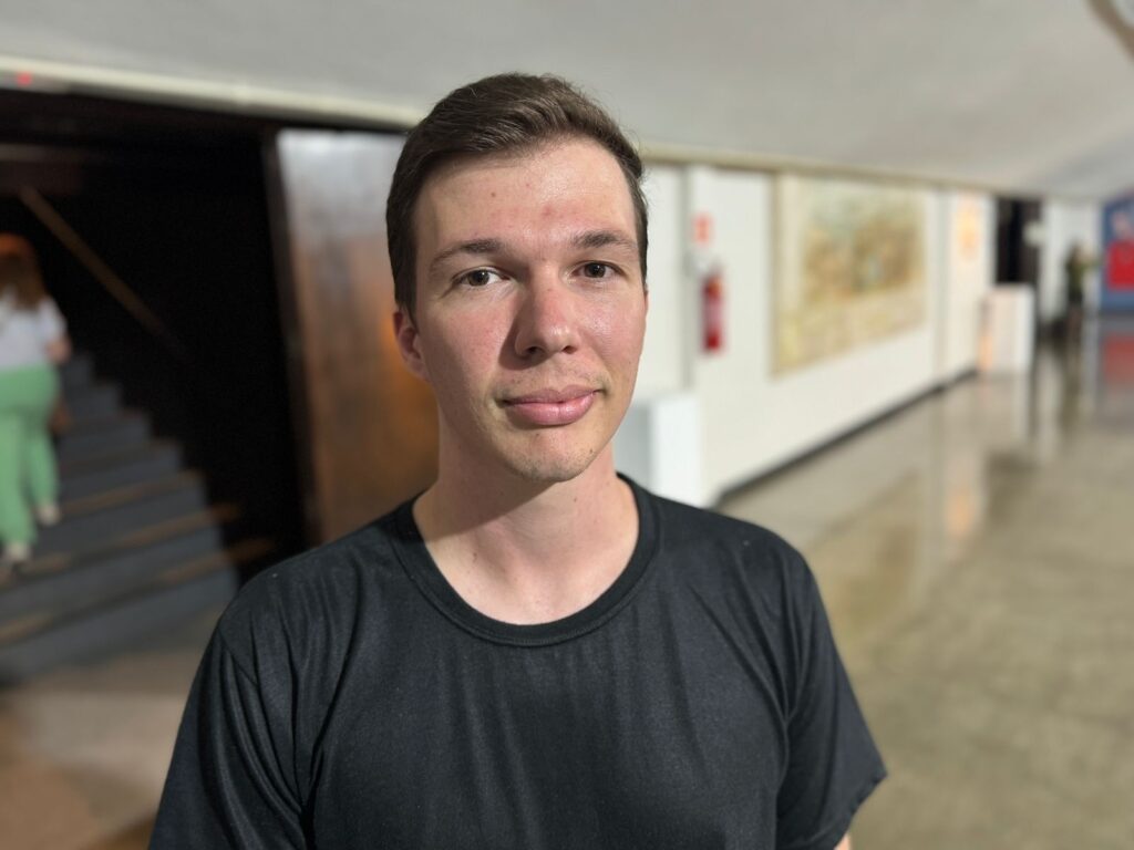 A imagem mostra um close-up de um jovem homem de pele clara, com cabelo castanho curto e liso. Ele está vestindo uma camiseta preta de gola redonda. Seus olhos são castanhos e ele tem uma expressão neutra no rosto. A iluminação é suave, lançando uma sombra sutil em seu rosto. Ao fundo, um corredor com paredes brancas e obras de arte penduradas. Há também uma escadaria escura à esquerda, com uma pessoa vestindo roupas verdes subindo. O chão é de concreto polido, refletindo a luz ambiente. A imagem tem uma profundidade de campo rasa, mantendo o foco no homem e desfocando o fundo.