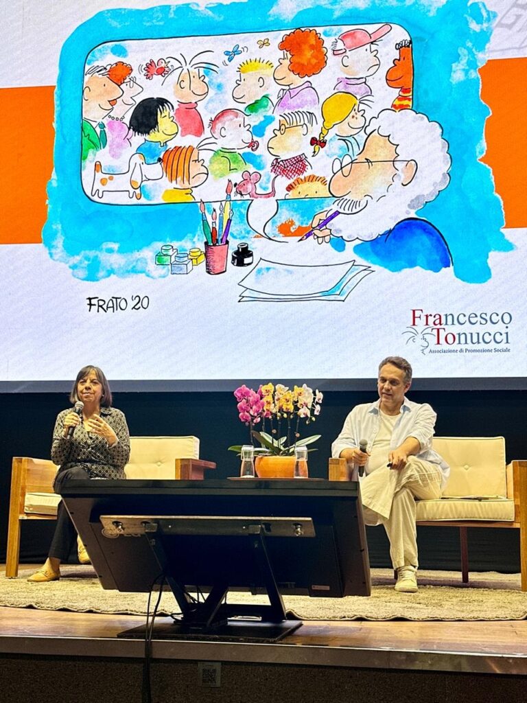 A imagem mostra um evento ao vivo com um painel de discussão. No palco, duas pessoas estão sentadas em poltronas de madeira, com uma mesa à frente delas. Uma mulher à esquerda está falando em um microfone, com as mãos gesticulando. Um homem à direita está sentado com um microfone na mão, olhando para a esquerda. Atrás deles, há um arranjo de flores e vasos. Acima do palco, um grande telão exibe uma ilustração colorida de um artista desenhando um grupo de personagens caricatos, que parecem crianças e adultos. A ilustração é assinada "FRATO '20" e, ao lado direito, há um logo com o nome "Francesco Tonucci" e "Associazione di Promozione Sociale". O fundo geral da imagem é escuro, com a iluminação focada no palco e no telão.