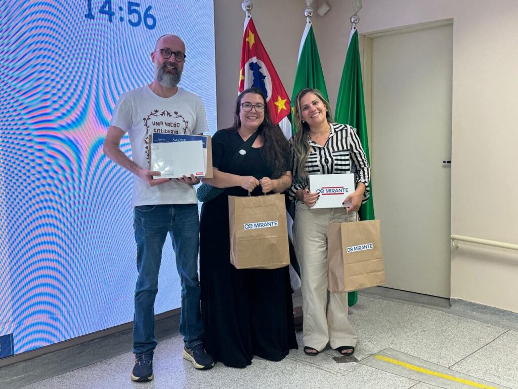 A imagem mostra três pessoas em pé, duas mulheres e um homem, em um ambiente interno com bandeiras ao fundo. O homem, à esquerda, veste uma camiseta branca com estampa, óculos e jeans. Ele segura uma caixa branca. Ao lado dele, uma mulher com óculos e um vestido preto longo segura uma sacola de papel marrom. A mulher à direita, com uma blusa listrada e calças claras, segura uma caixa branca e outra sacola de papel marrom. Todas as sacolas de papel e uma das caixas têm o logo "MIRANTE" estampado. Atrás deles, há bandeiras verdes e uma bandeira com as cores da bandeira do Brasil e uma estrela amarela. Ao fundo, à esquerda, um grande painel digital exibe o horário "14:56".