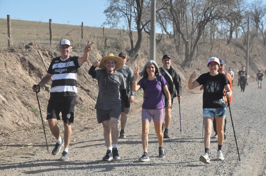 A imagem mostra um grupo de pessoas caminhando em uma estrada de terra em um dia ensolarado. A maioria dos indivíduos está usando roupas casuais e parece estar aproveitando a atividade ao ar livre. Alguns deles estão usando bastões de caminhada, o que sugere que eles podem estar participando de uma trilha ou caminhada mais longa. No primeiro plano, um homem com uma camisa listrada em preto e branco e shorts pretos está posando para a câmera com um sorriso e um gesto de paz. Ao lado dele, um homem com um chapéu de palha e uma camiseta cinza também está sorrindo e fazendo um gesto semelhante. Mais atrás, outras pessoas caminham, com algumas delas também fazendo gestos para a câmera. O cenário é composto por uma encosta gramada e algumas árvores secas ao fundo, com um céu azul claro acima.
