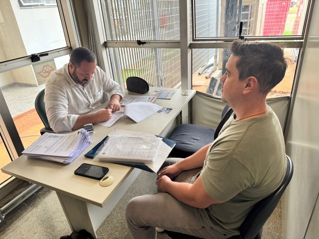 A imagem mostra dois homens sentados em uma sala, possivelmente um escritório. O homem à esquerda, vestindo uma camisa branca, está sentado à mesa e escrevendo em um documento. Há várias pilhas de papéis e um celular sobre a mesa. O homem à direita, vestindo uma camiseta verde, está sentado em uma cadeira e olhando para o homem à esquerda. A sala tem janelas grandes que deixam entrar muita luz natural. O exterior das janelas mostra uma área com grades e algumas construções.